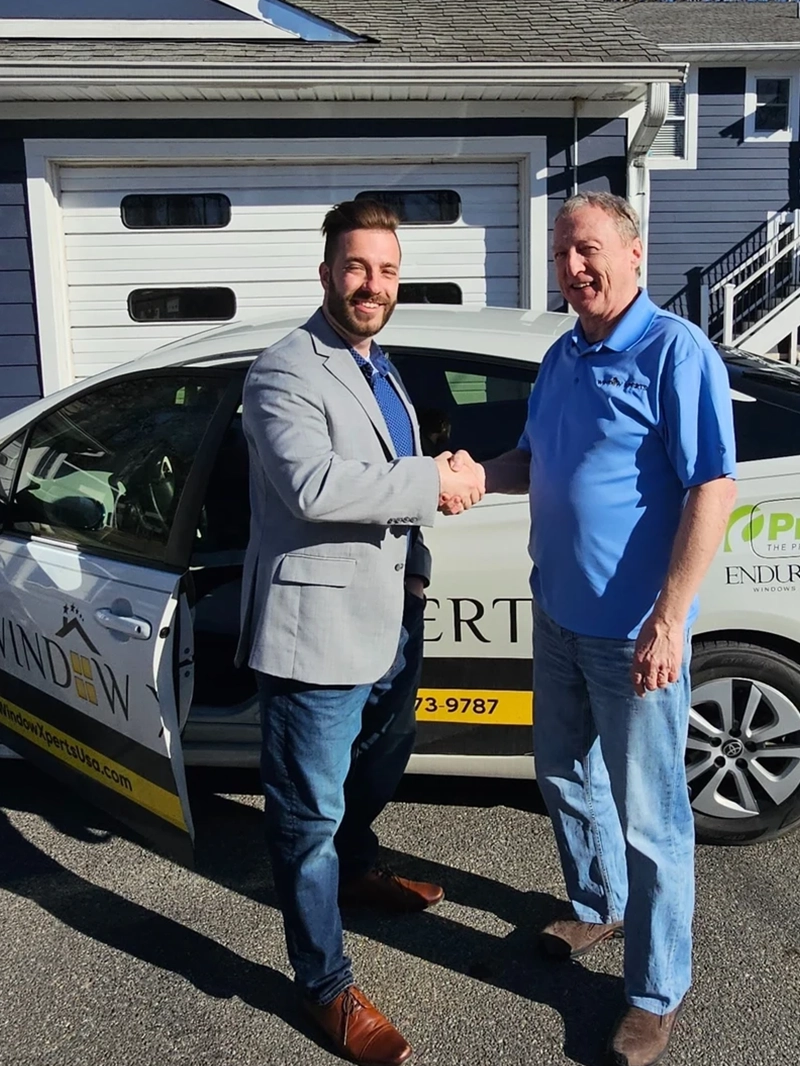 Two men shake hands beside a parked car with a logo, set against a suburban house and blue sky.